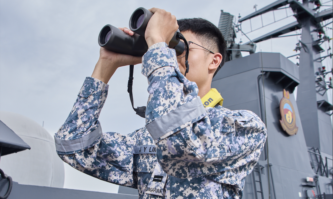 Naval officer holds a binoculars on a warship’s deck under blue sky.